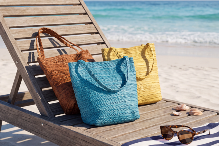 Three woven tote bags in brown, blue, and yellow are arranged on a wooden beach lounge chair with a sandy beach and ocean in the background.